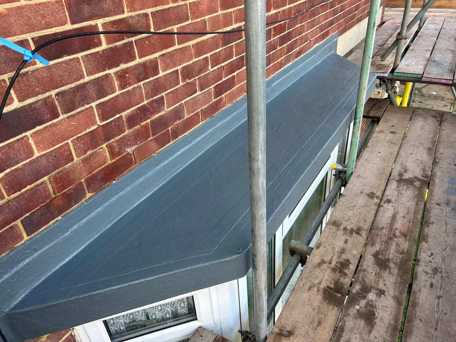 Gray painted awning above a window on a red brick wall next to wooden scaffolding planks.