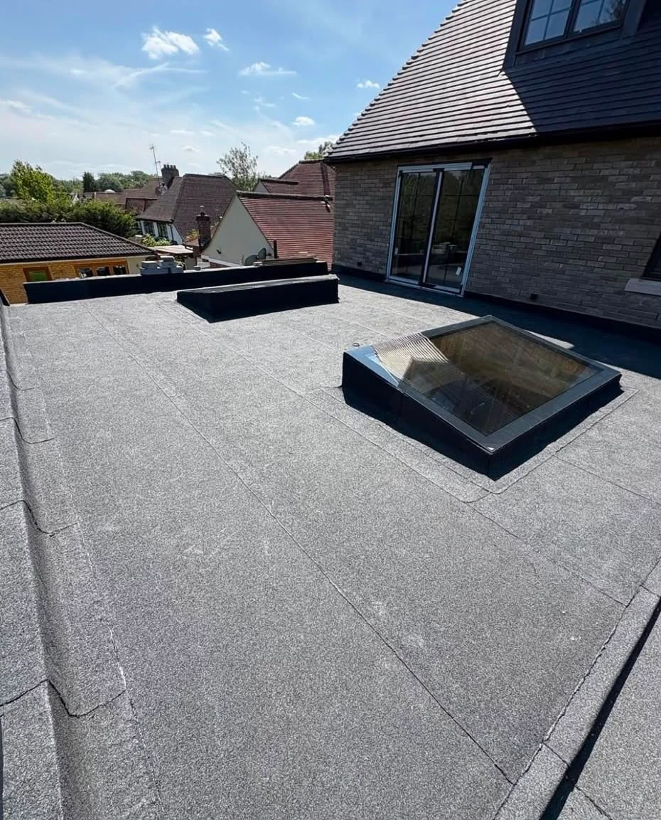 Flat rooftop with a glass skylight and nearby houses under a partly cloudy blue sky.