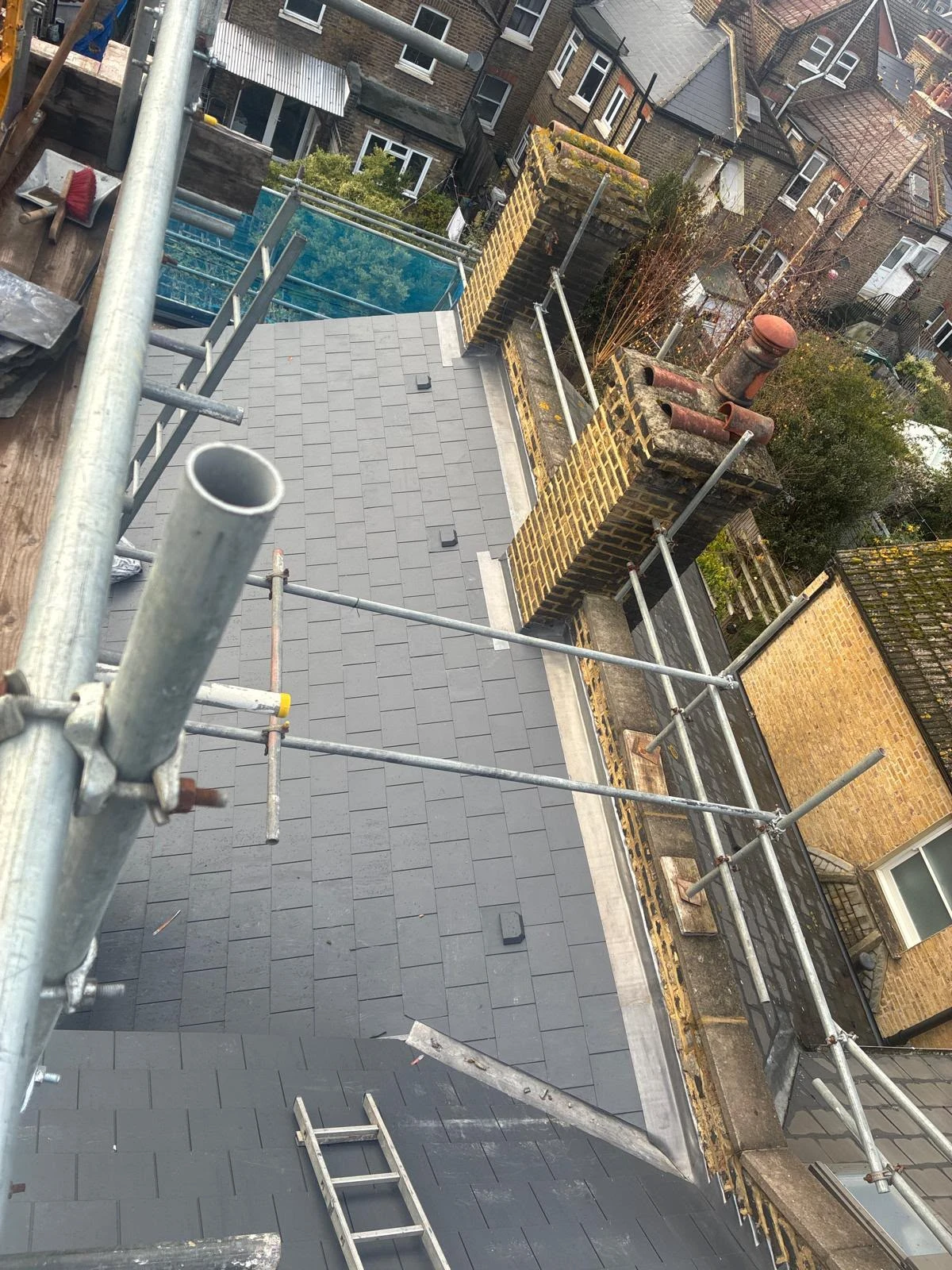 View of a rooftop with new gray tiles, metal scaffolding, chimney stacks, and nearby brick buildings.