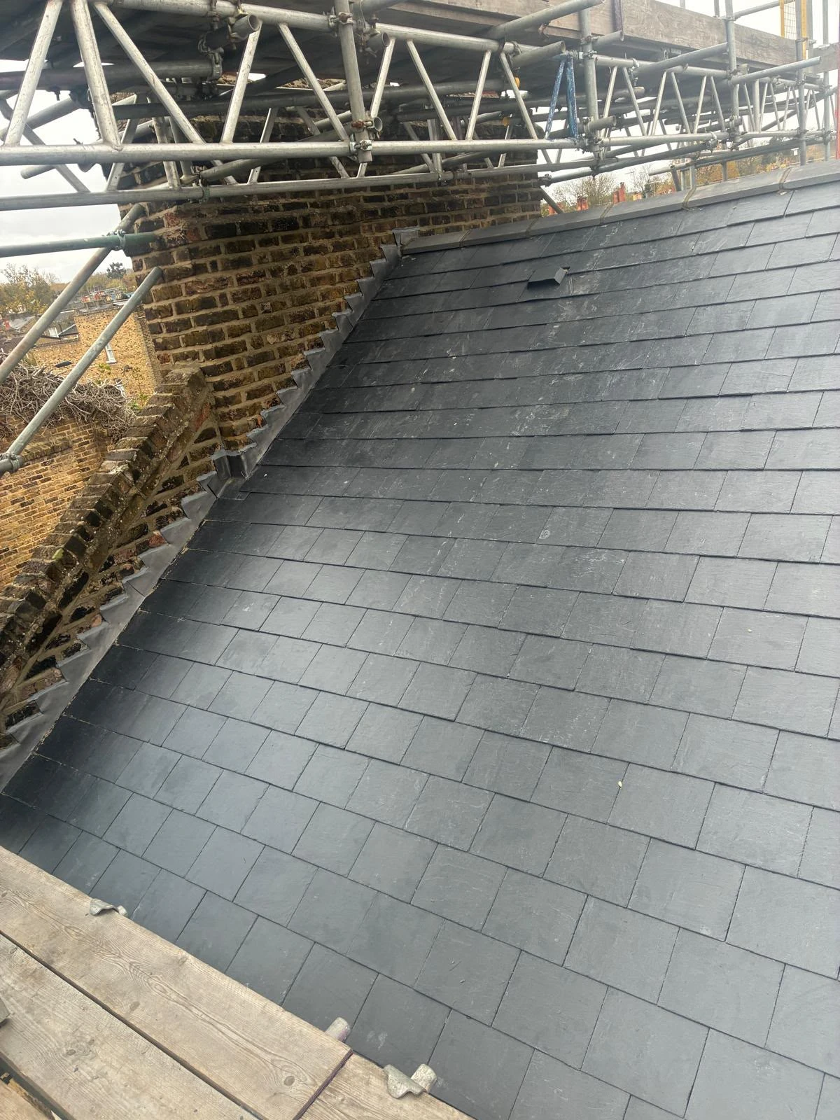 Slate tiled roof section next to a brick chimney, with metal scaffolding above and wooden planks below.