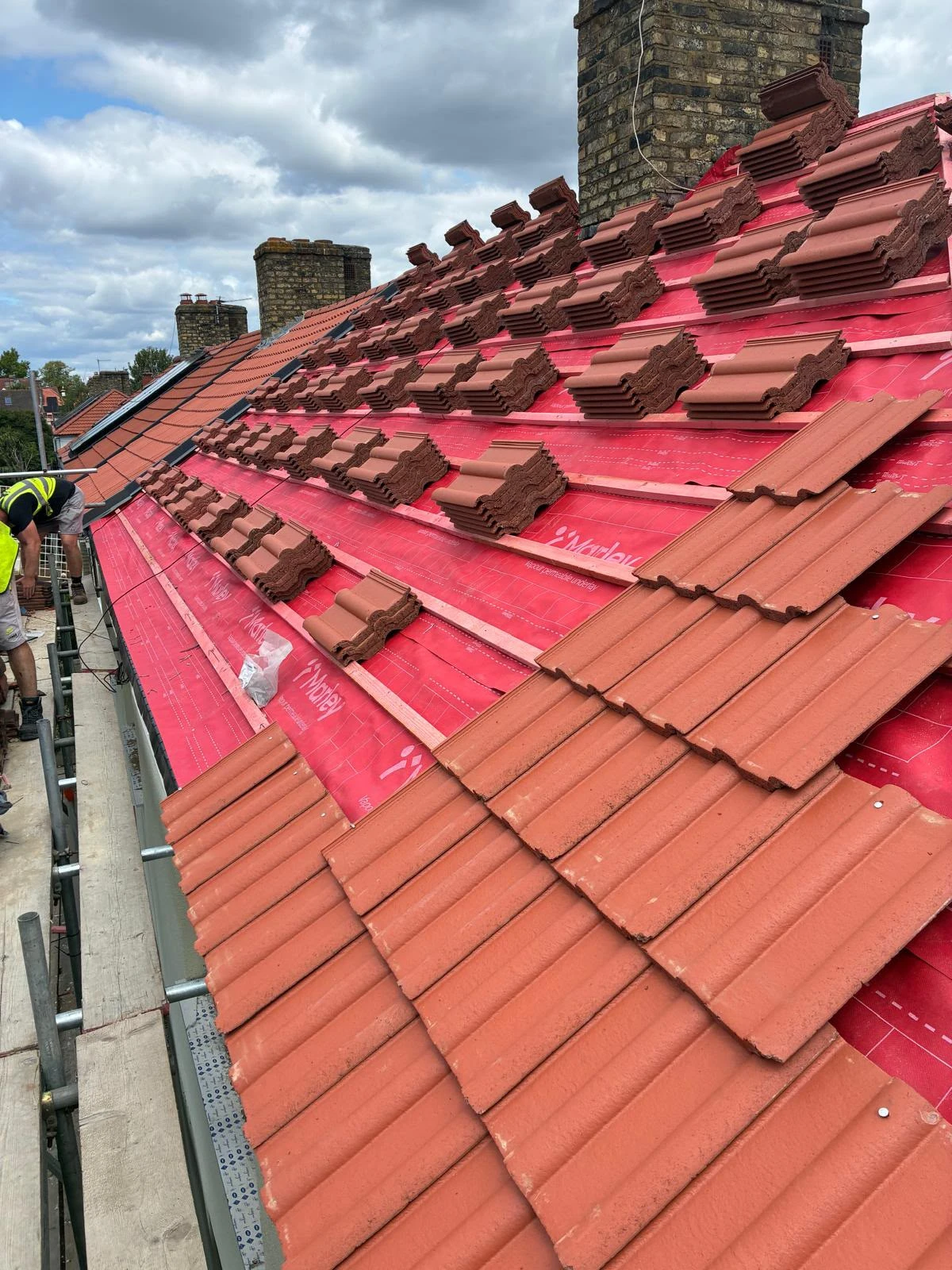Roof under construction with red tiles stacked on red underlayment and a worker on scaffolding.