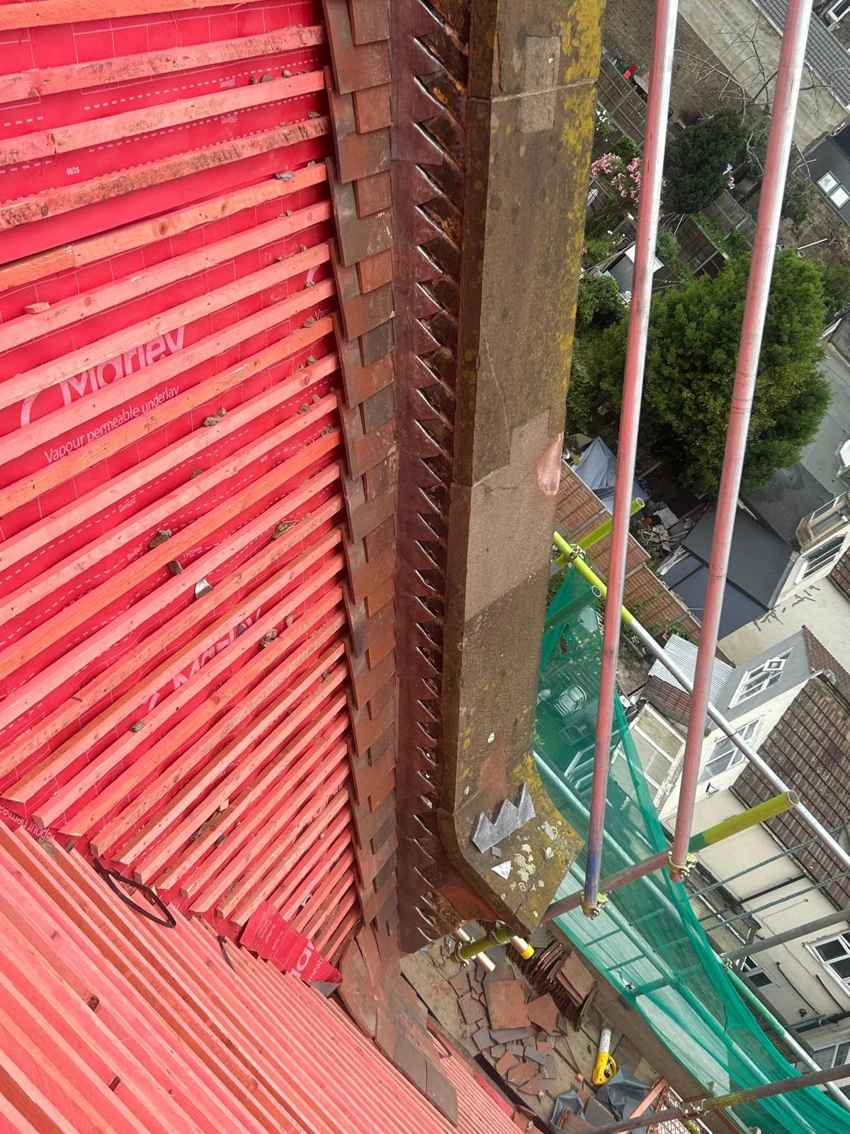 View of a roof under construction with red wooden battens, old tiles, and scaffolding outside a house.