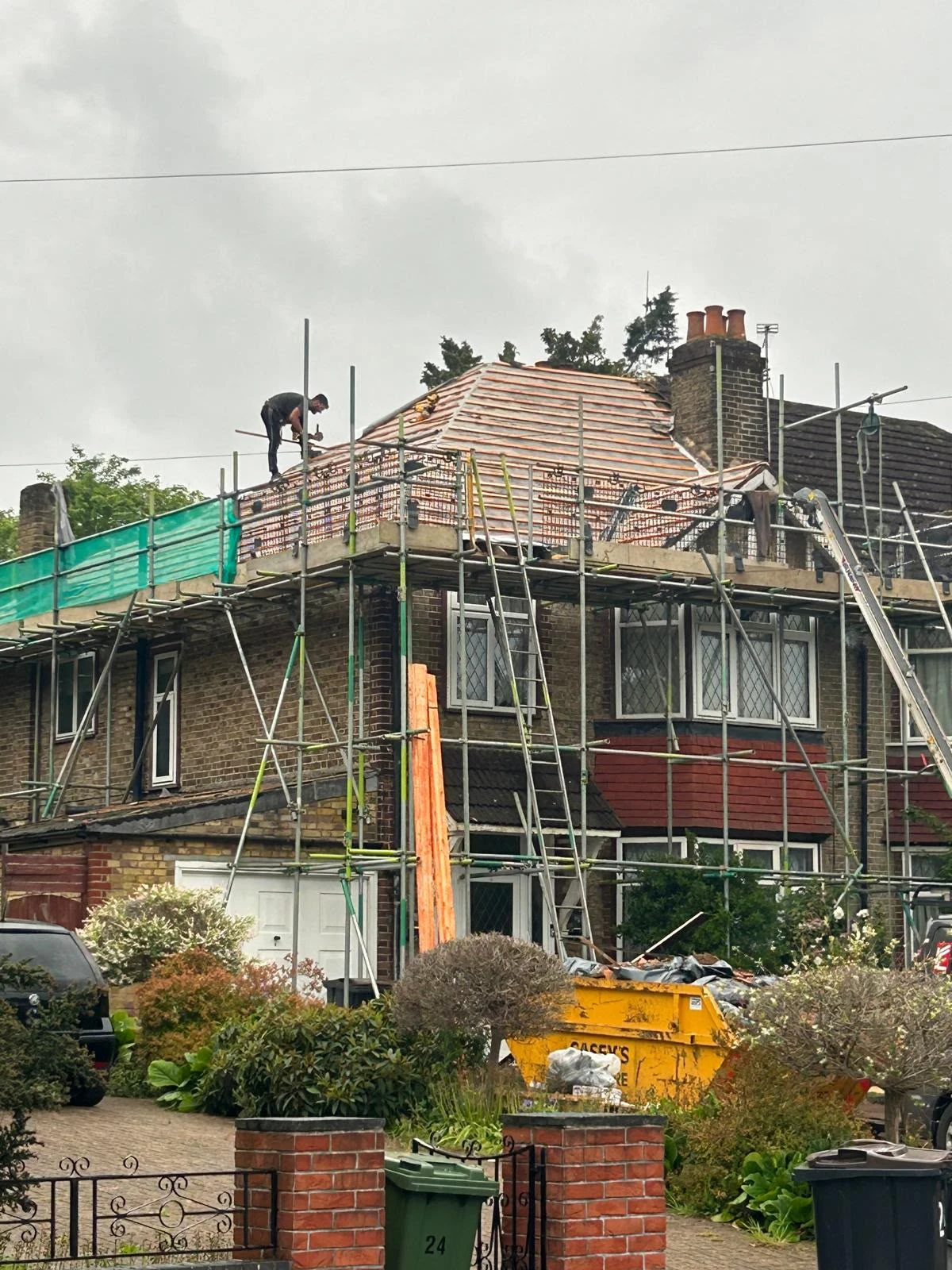 Worker on scaffolding repairing roof of a brick house with construction materials and bins in front yard.