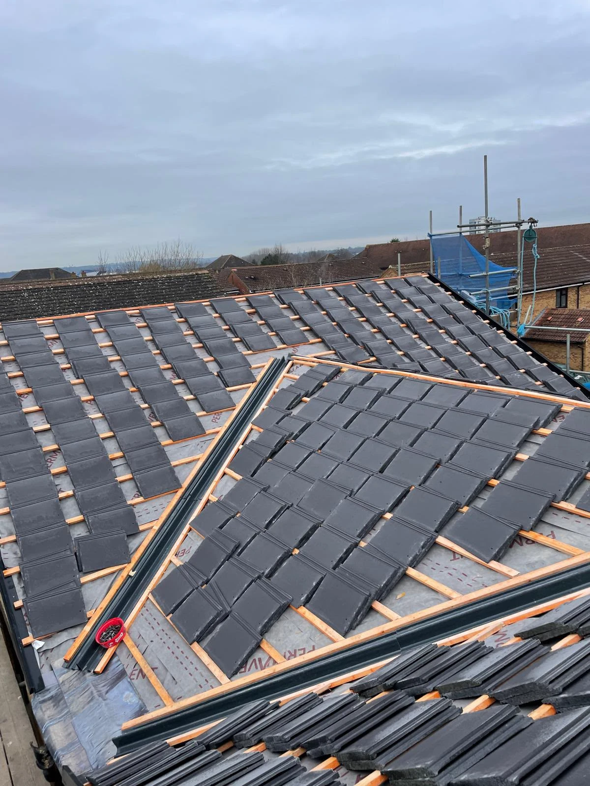Partially tiled roof with black tiles laid out on wooden battens under cloudy sky.