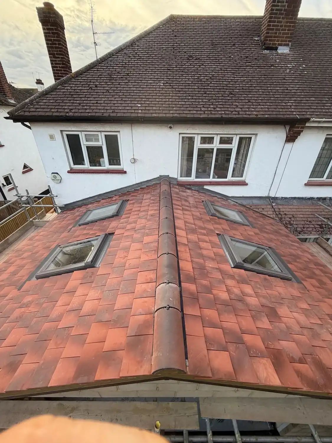 View of a red tiled roof with four skylight windows, centered ridge tiles, and a larger house with white walls and brown tiled roof in the background.