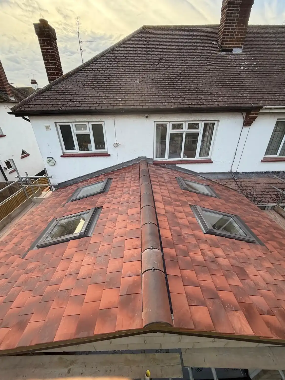 Red tiled roof extension with four skylight windows, two on each side of a central ridge, attached to a white house with brick chimneys and multiple windows on the upper floor.
