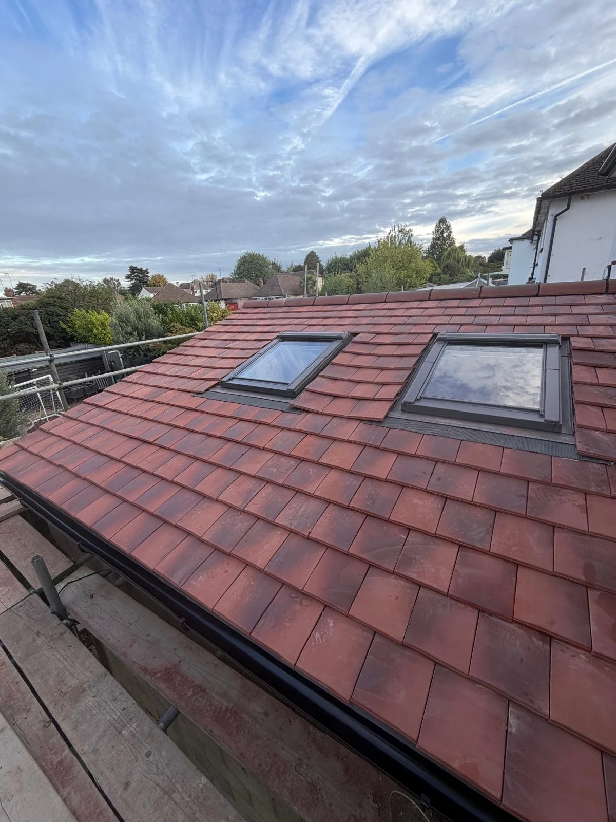 Sloped roof with red clay tiles featuring two rectangular skylight windows under a partly cloudy sky in a suburban area with trees and houses nearby.