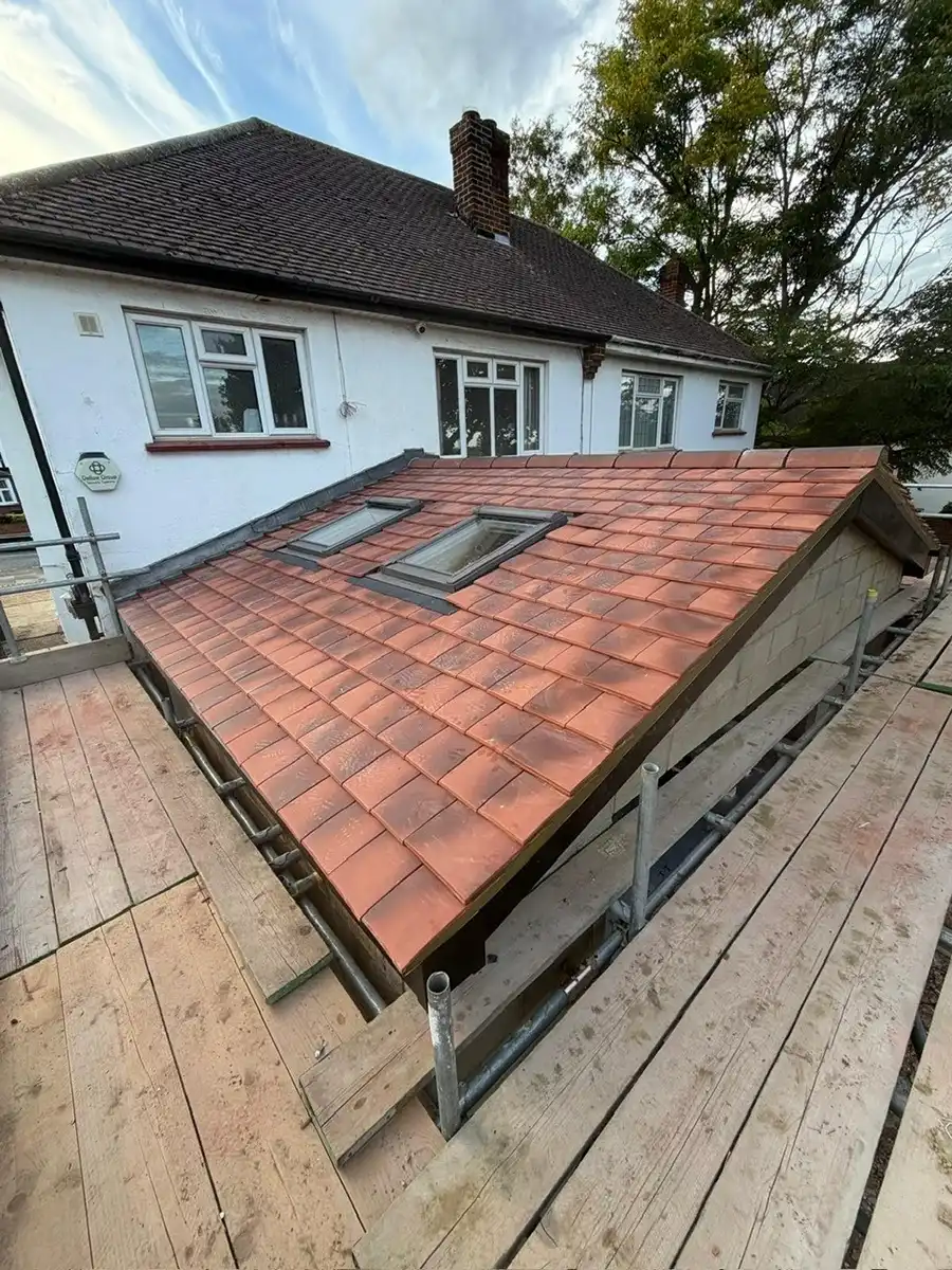 Newly tiled red roof extension with two skylight windows next to a white two-story house with brown chimneys, surrounded by wooden scaffolding.