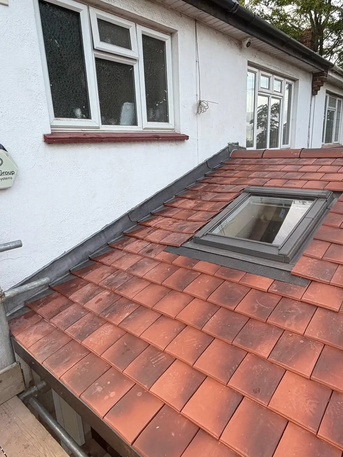 Close-up of a red clay tiled roof with a rectangular skylight window installing next to a white exterior wall with two windows and a drainpipe.