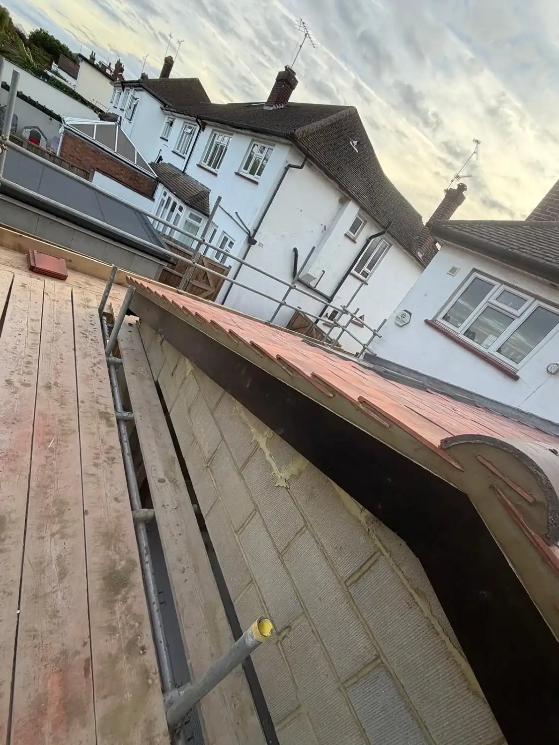 View from a scaffold platform showing a section of a building with newly installed reddish roof tiles, a concrete block wall, and neighboring white houses with pitched roofs under a cloudy sky.